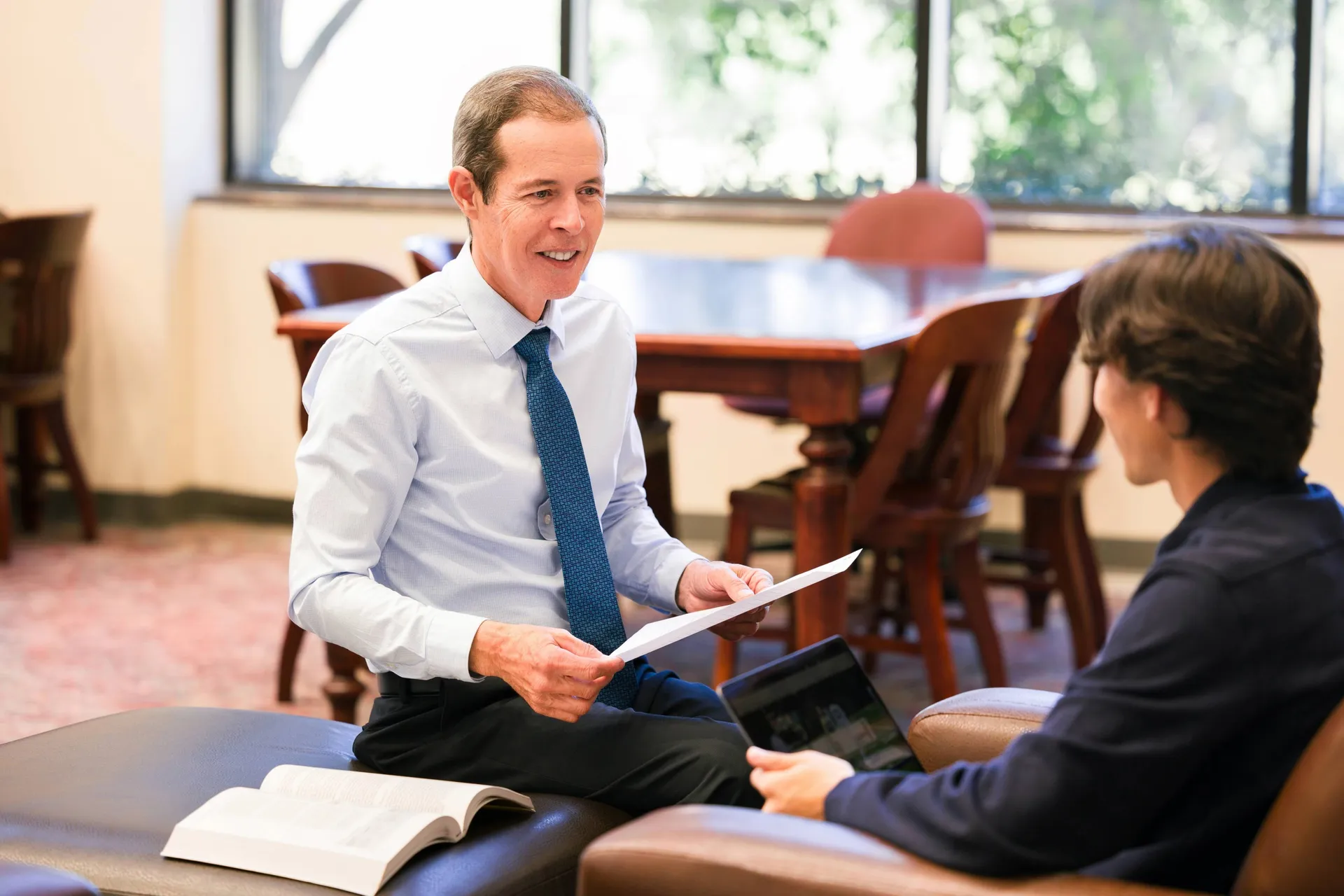 A teacher holding a paper talking to a student sitting in a chair with a laptop