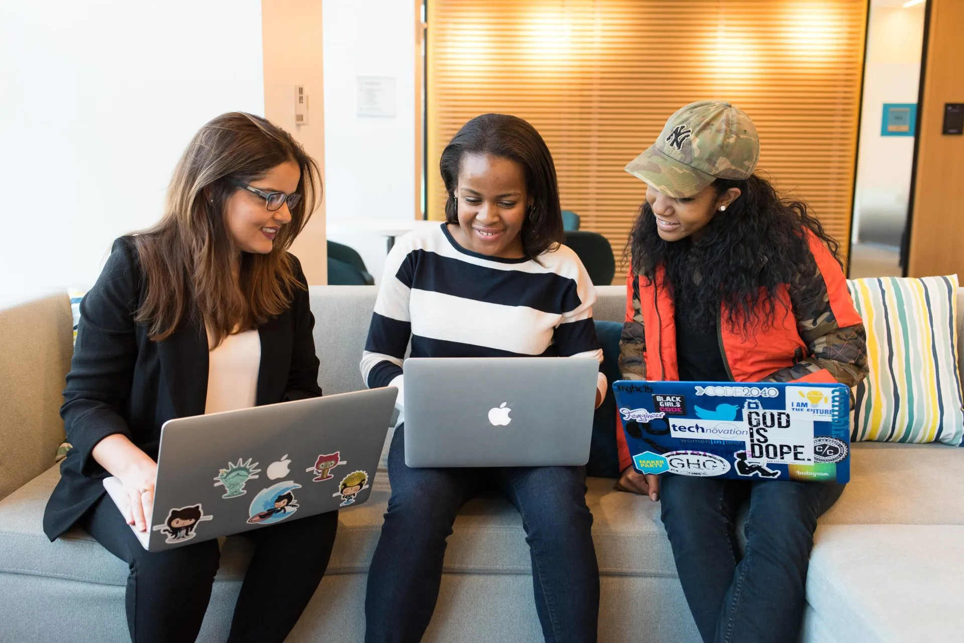 Three women sit on a couch with laptops in their lap, looking at each other's screens