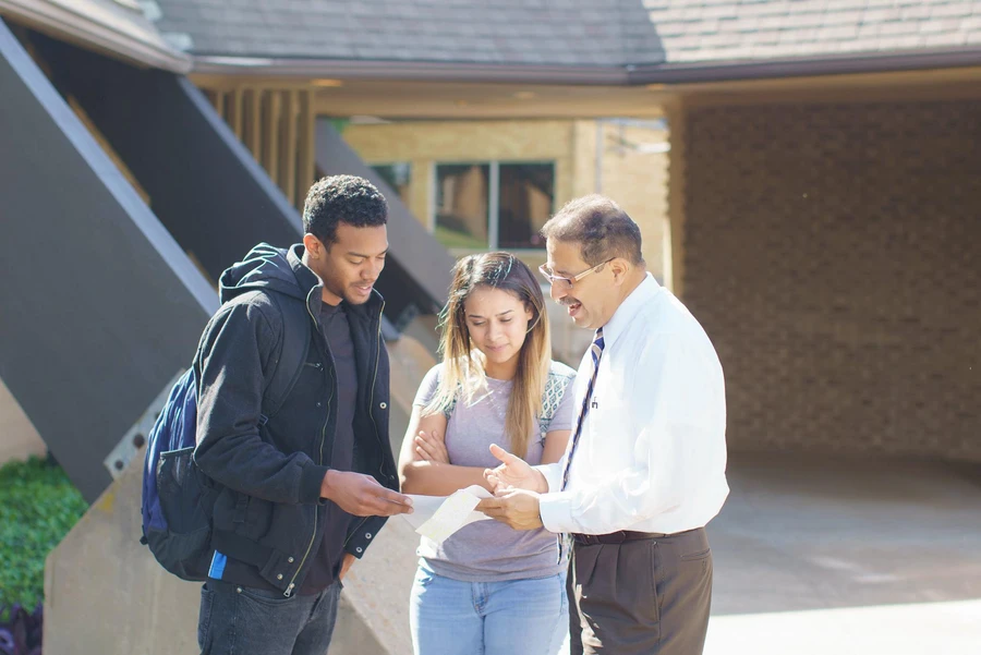 A female and a male student talking with Professor Rico, in front of Barron Chapel, as they read a note card. 