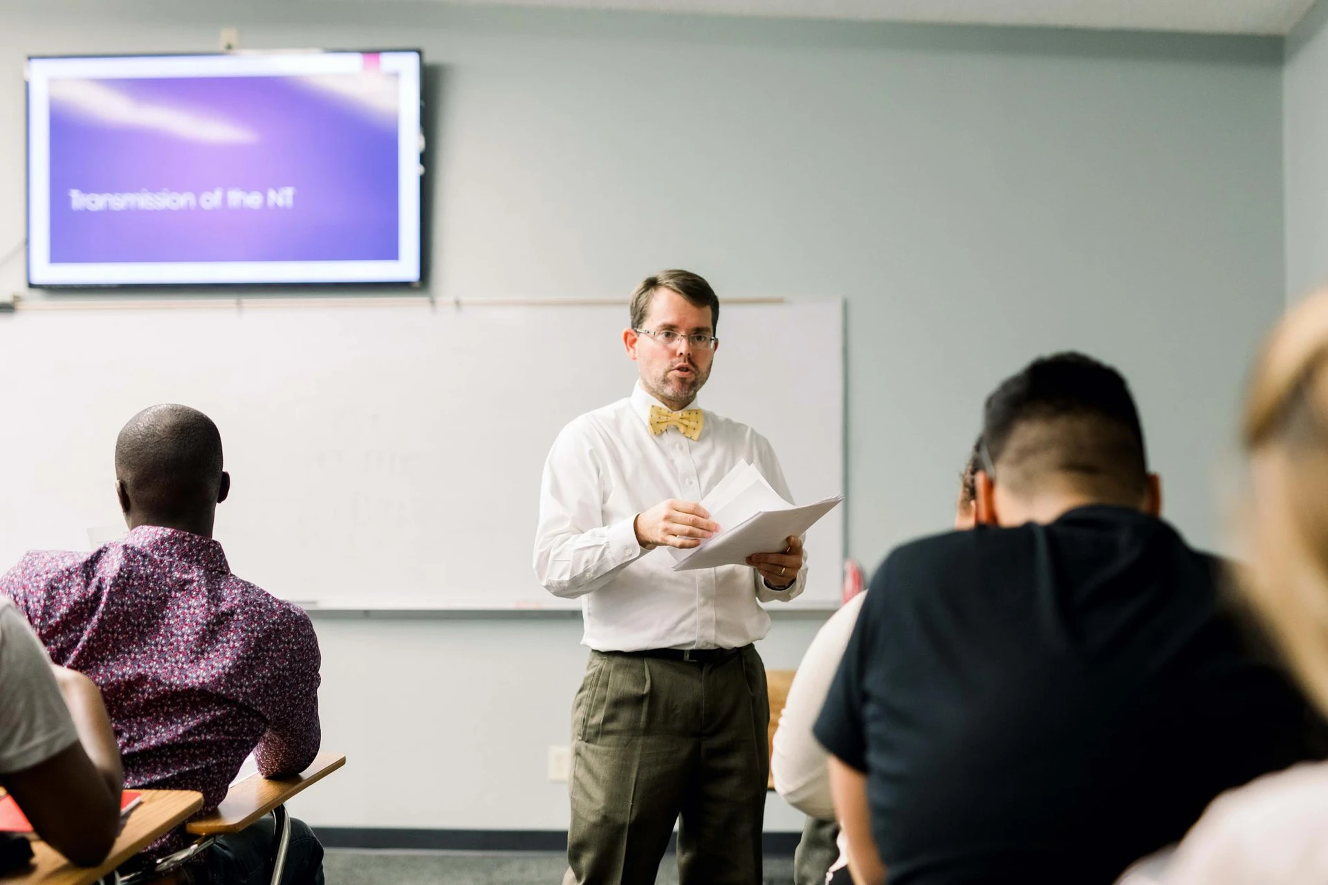Dr. Campbell, dressed in a white buttoned shirt and yellow bow-tie, stands in front of his class and holds papers as he begins to hand them out to his students