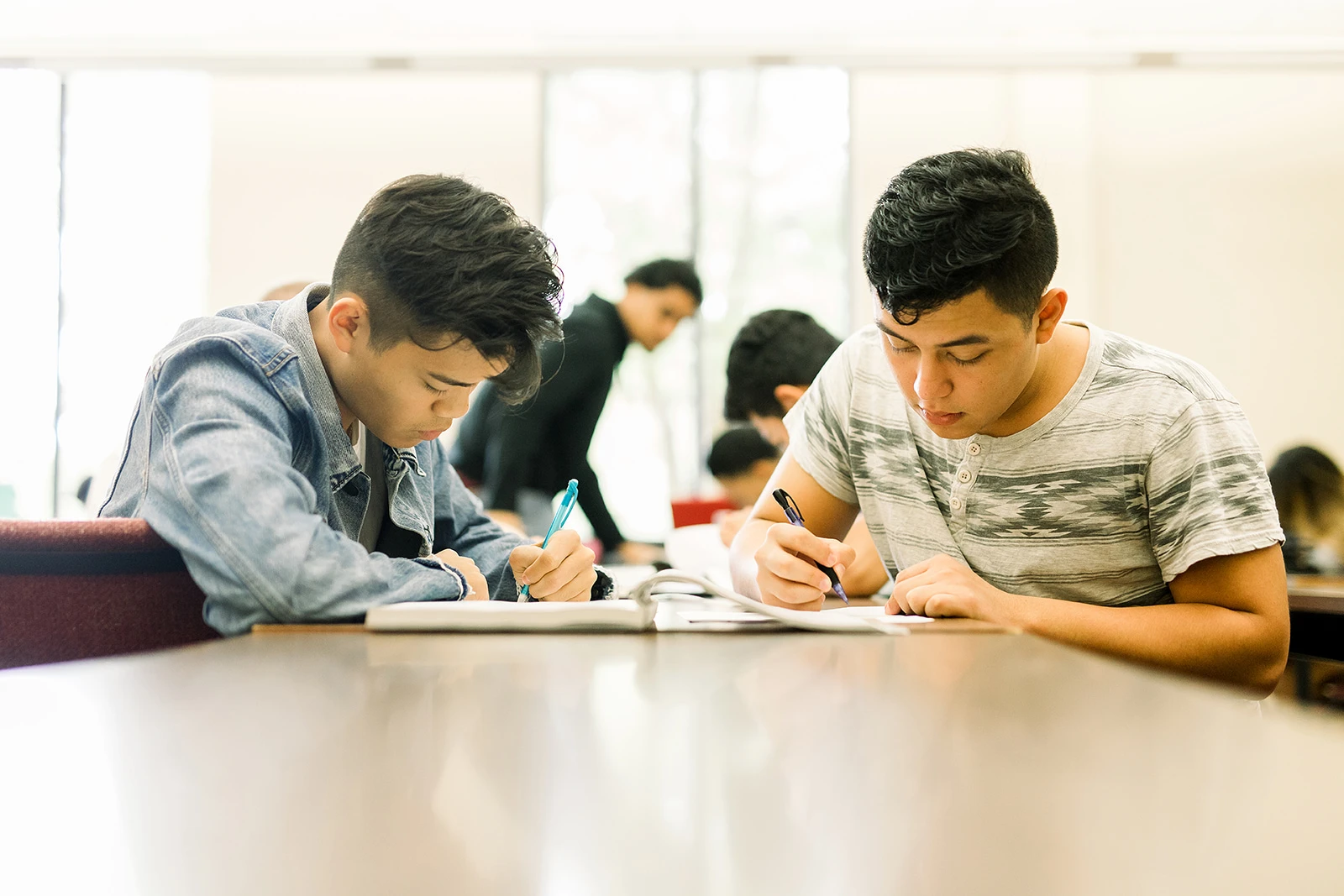 Two male students sit at a rectangle table taking notes from the same textbook