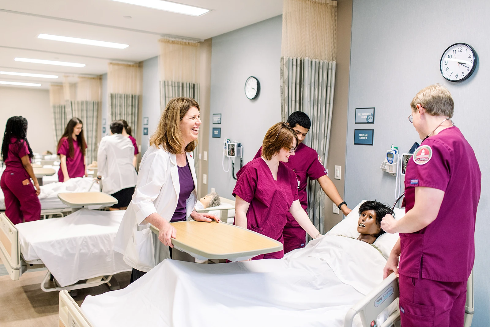 Three nursing students dressed in maroon scrubs look down at their patient and their professor watches over and smiles