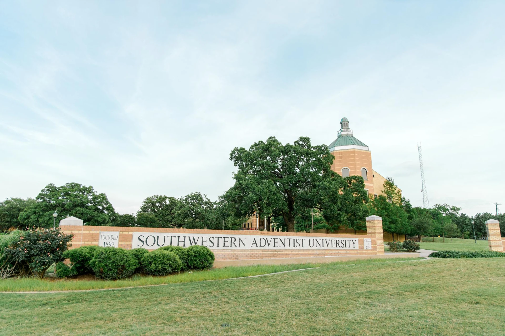 Campus sign made of bricks that reads "Southwestern Adventist University" with the top of Pechero Hall showing behind trees