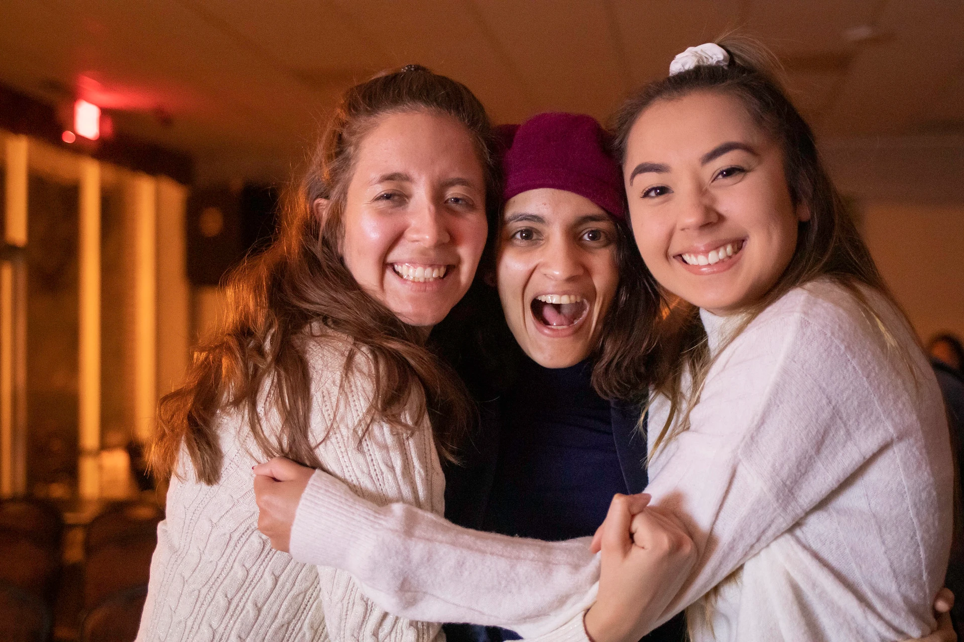 Three female friends hug tightly and smile as they are dressed in long-sleeved sweaters
