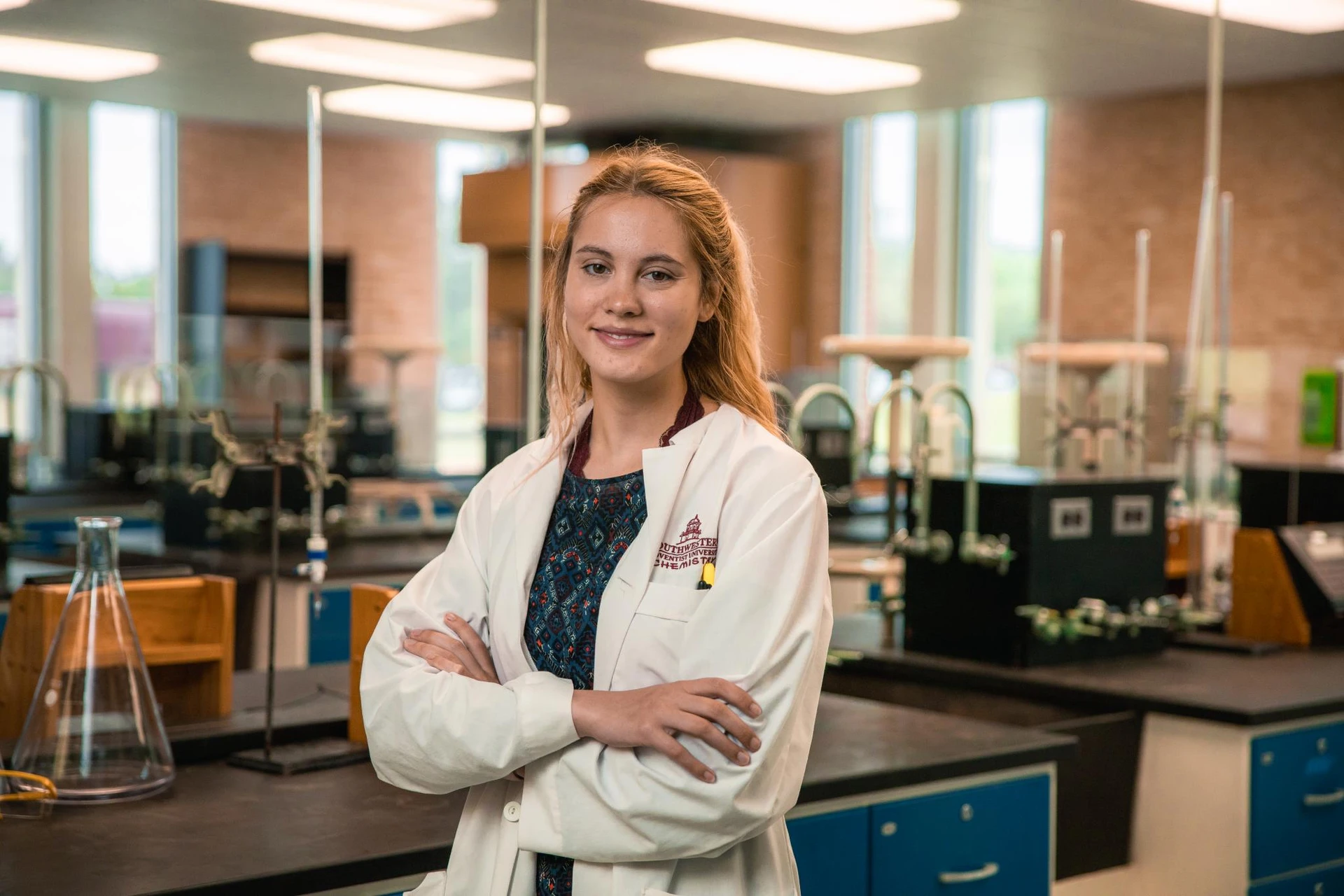 Kelsey Johnson poses in the lab with her arms crossed wearing a lab coat that reads "Southwestern Adventist University Chemistry"