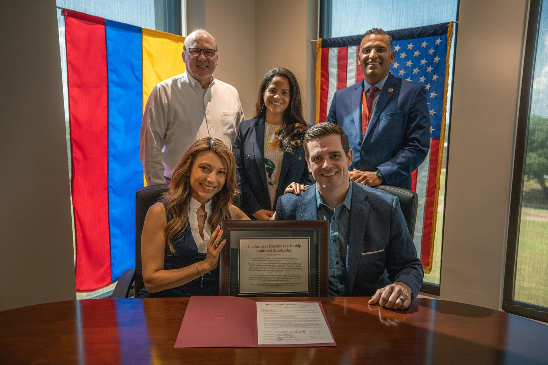 The Vallettas pose holding up a frame with Joel Wallace, Ana Patterson, and Tony Reyes standing behind them with the Columbian and American flags behind them.