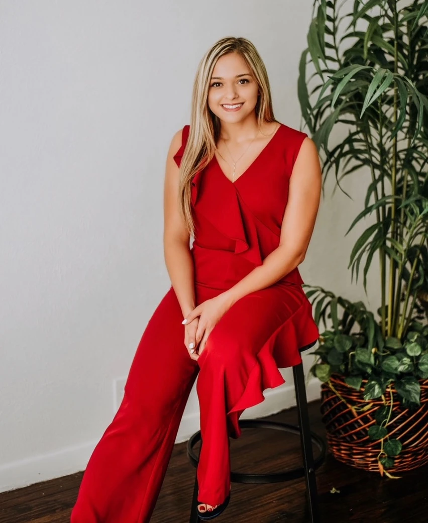 Anabella Lopez poses sitting on a chair next to a plant in a sleeveless red jumpsuit