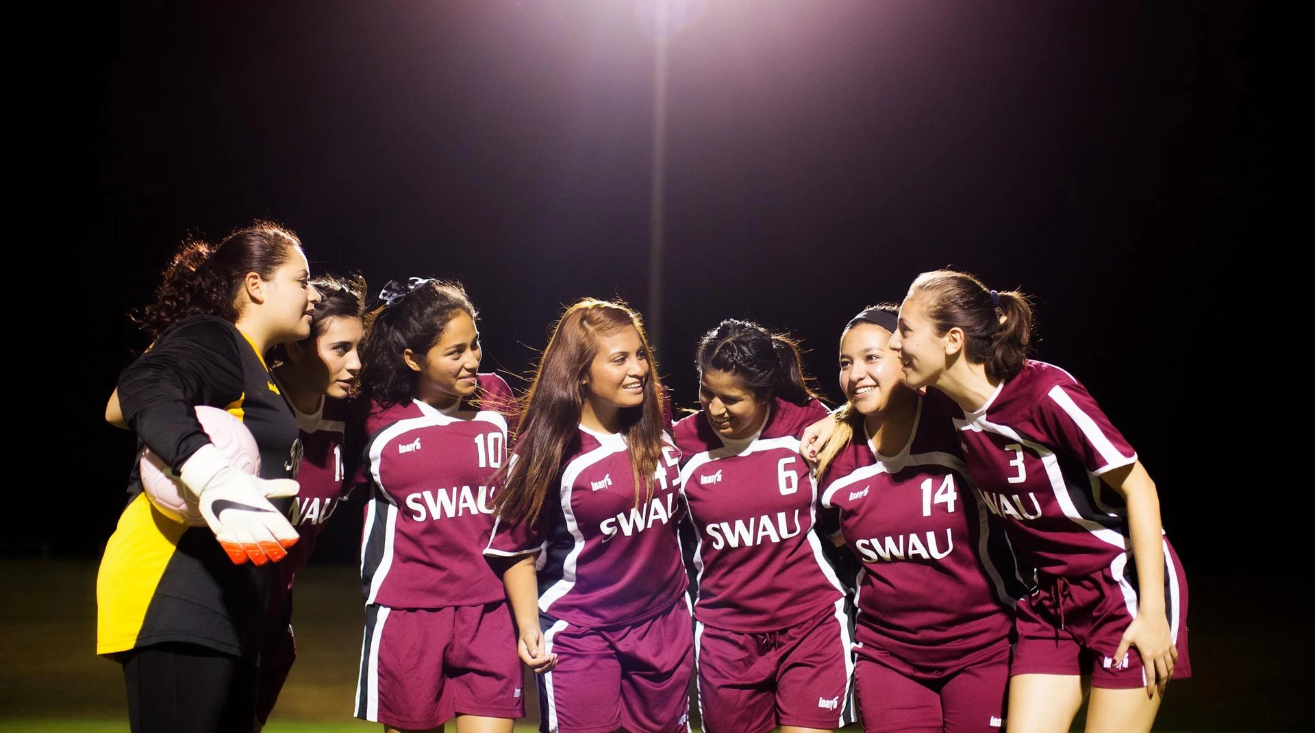 A group of female soccer players wearing maroon SWAU jerseys stand in a semi circle and talk