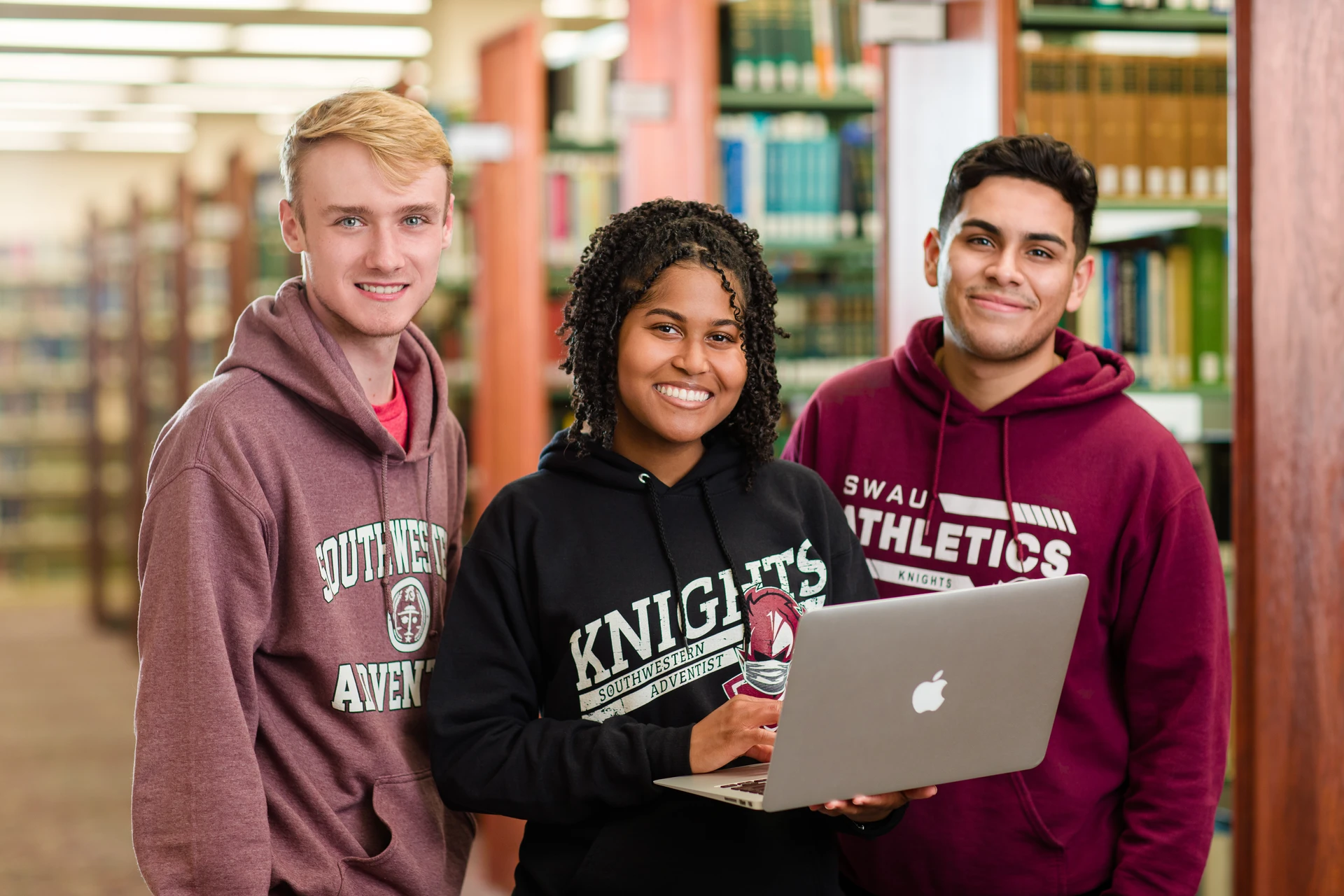 Three Adventist University students smile at the camera standing in a library