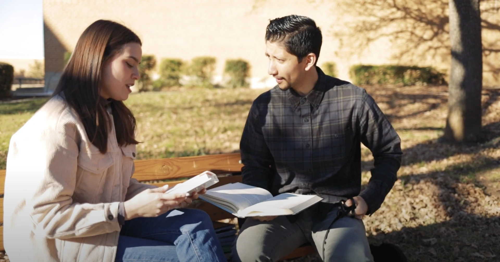 Two students sit on a bench holding books and talking