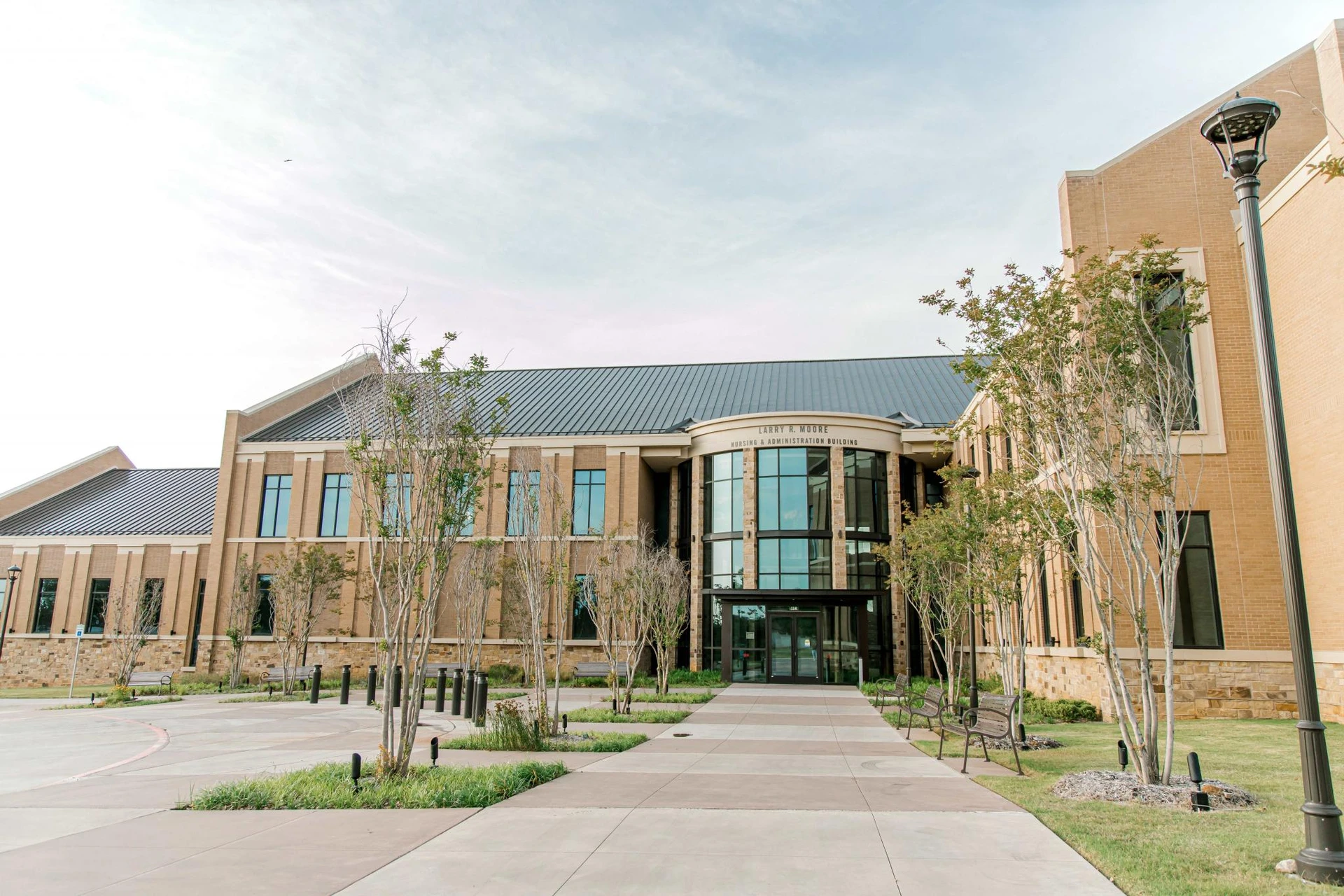 A wide sidewalk, with small trees and benches on the side, lead up to the doors of the new nursing and administration building named Larry R. Moore.