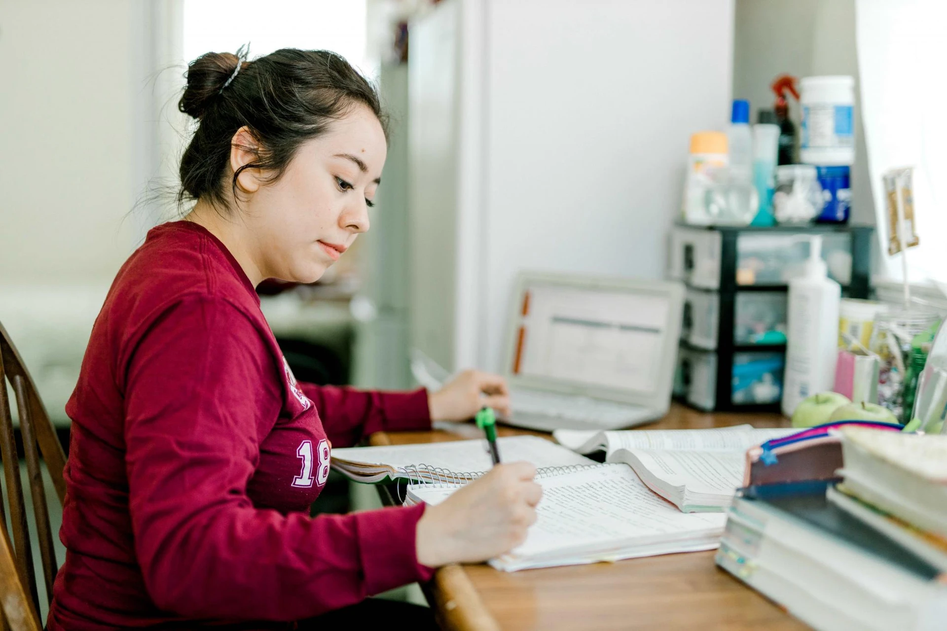 A student, dressed in a maroon long-sleeved shirt and with her hair in a bun, sits in front of a table as she completes her homework 