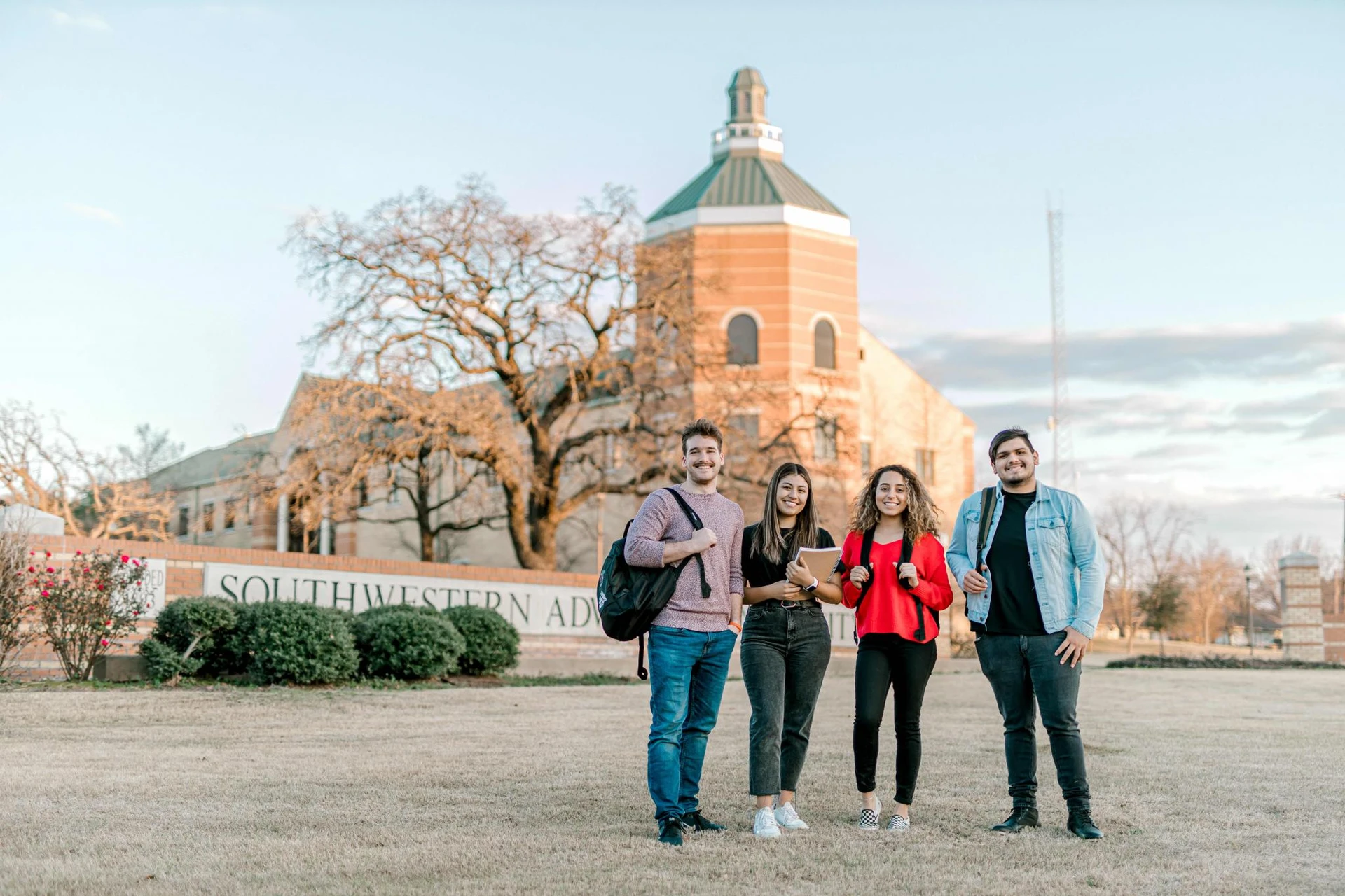 Four students, with backpacks, smile together as they stand in front of a bricked sign that reads " Southwestern Adventist University" 