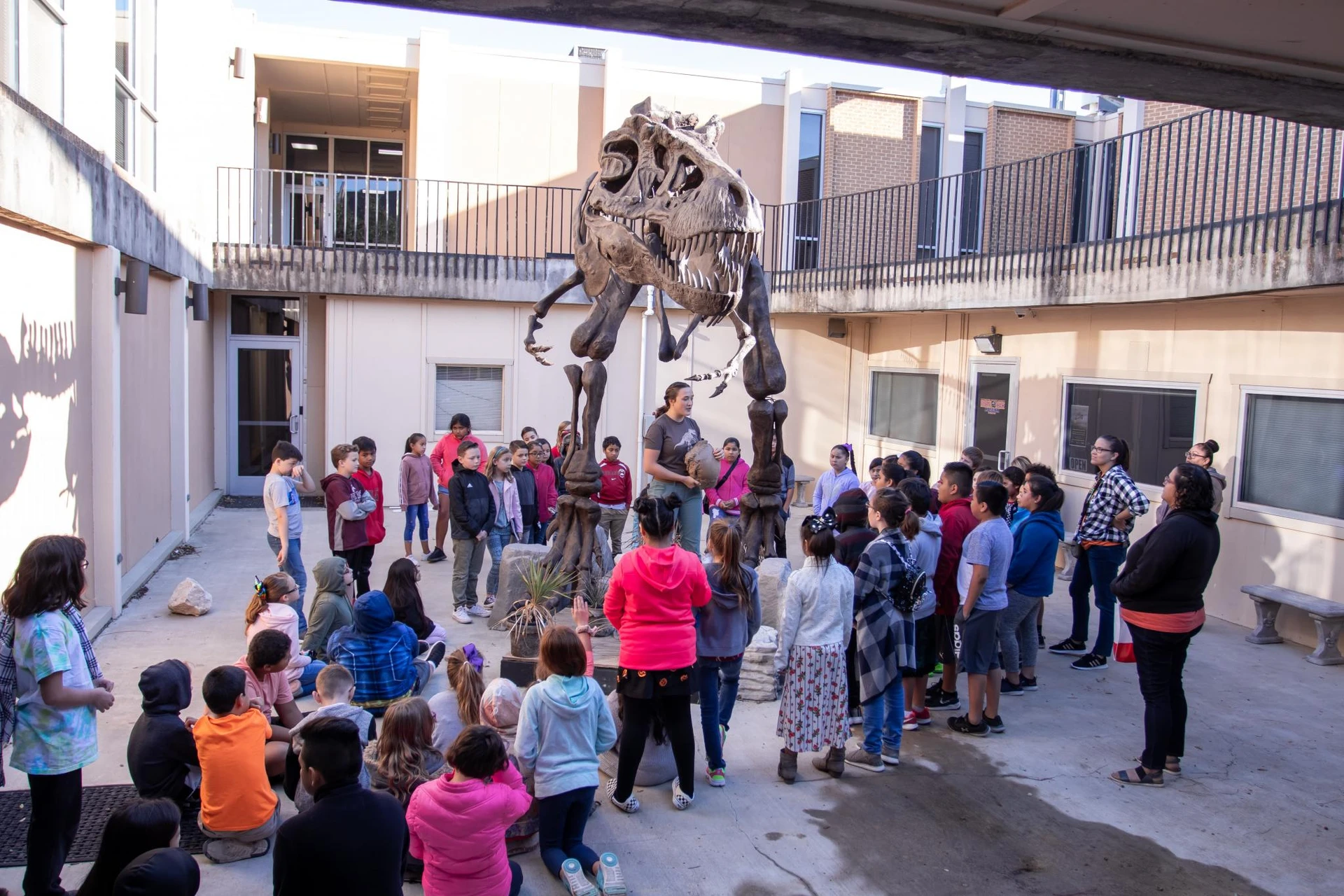 Kids surrounding the T-Rex skeleton look forward and listen to the dinosaur museum worker explain something